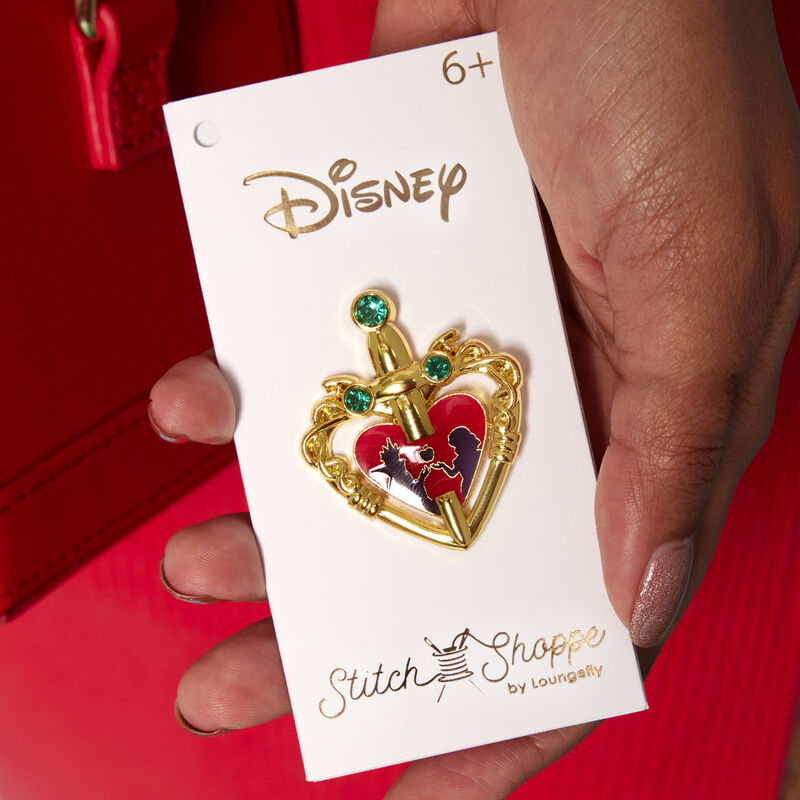 Hand holding the pin that comes with the Evil Queen Heart Box Crossbody Bag, featuring a gold heart with a sword through it that has the image of Snow White and the Evil Queen's silhouettes in the center